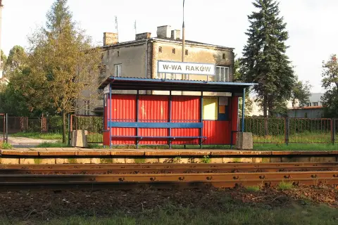 Przystanek kolejowy Warszawa Raków. Czerwona wiata z niebieską ławką i tablicą informacyjną. W tle budynek i wysokie drzewa.