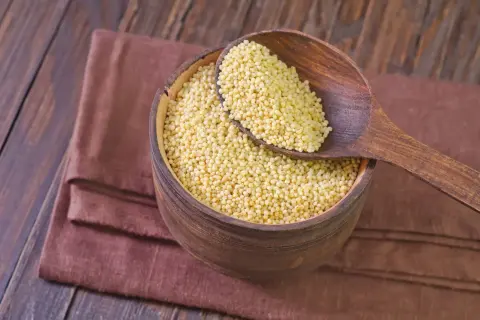Drewniana miska pełna kaszy jaglanej, z łyżką nabierającą jej ziarna. Czy kasza jaglana ma gluten? Nie, jest bezglutenowa!