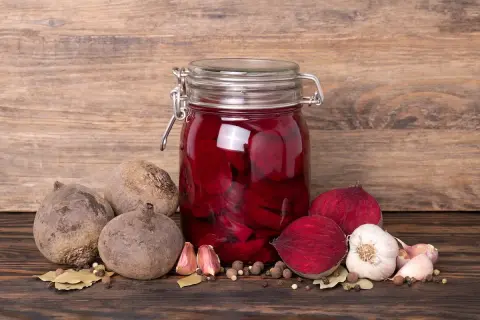 Ein Glas mit eingelegten, roten Bete Scheiben steht auf einem Holztisch. Daneben liegen frische rote Bete, Knoblauch und Gewürze. Das Bild zeigt den Prozess des rote Bete fermentieren.