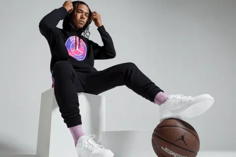 Mężczyzna w czarnej bluzie PSG Jordan z logo, czarnych spodniach dresowych, białych butach i różowych skarpetkach siedzi na białych sześcianach.