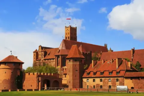 Potężny zamek w Malborku, ceglana twierdza z czerwonymi dachami, góruje nad zielonymi polami. Zdjęcia tego miejsca zachwycają.