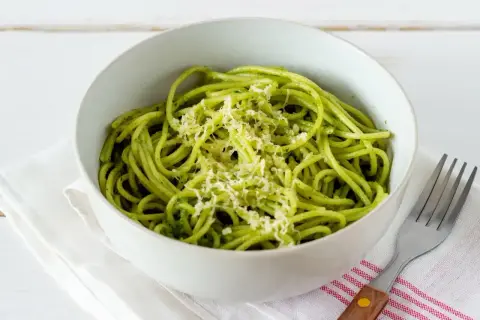 Miseczka spaghetti z zielonym, aromatycznym pesto z pokrzywy, posypana startym parmezanem. Obok leży widelec.
