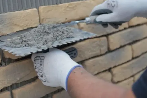 Ręce w rękawiczkach nakładają zaprawę na kamień dekoracyjny betonowy, przygotowując go do ułożenia na ścianie.