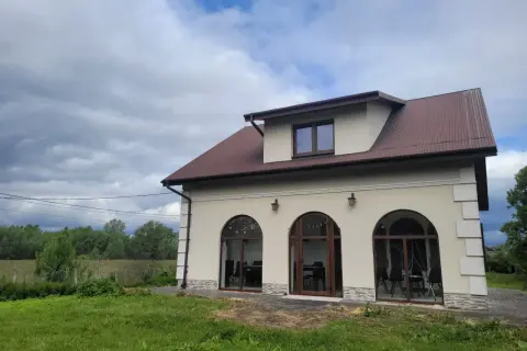Nowy dom w Charsznicy z łukowymi oknami i brązowym dachem, otoczony zielenią i chmurami na niebie.