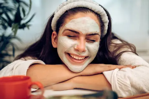 Lächelnde Frau mit Gesichtsmaske, die sich fragt: nach Gesichtsmaske eincremen oder nicht? Entspannter Moment mit rotem Becher.