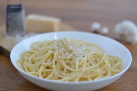 Ein Teller mit dampfenden Spaghetti, bestreut mit frisch geriebenem Parmesan. Dazu ein Stück Parmesan, eine Reibe und Knoblauch – perfekt für ein authentisches italienisches Gericht.