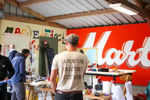 Des peintres travaillent dans un atelier, utilisant du matériel de peinture pour créer des lettres sur un mur. Le t-shirt d'un homme porte l'inscription "La peinture en lettres, c'est bien".