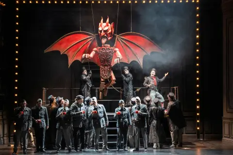 Bühnenszene der Oper Wuppertal: Eine Teufelsmarionette schwebt über Darstellern in Uniformen und Zylindern.