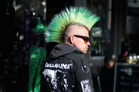 Mężczyzna z zielonym irokezem, w okularach przeciwsłonecznych i kurtce z naszywkami. Jego styl to czysty punk.
