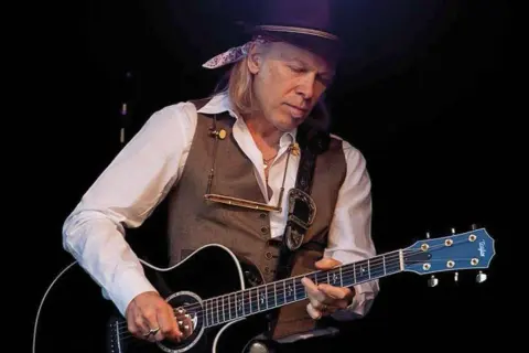 Elliott Murphy Zaragoza, con sombrero y chaleco, toca la guitarra acústica en un concierto.