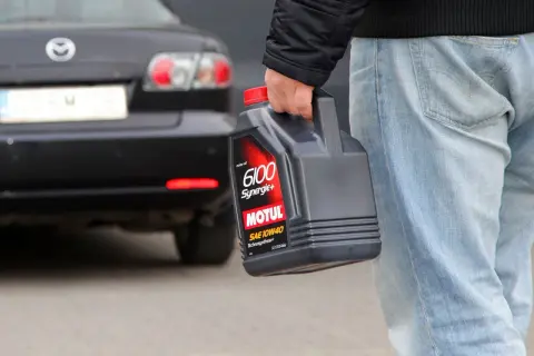 Mężczyzna niesie kanister z olejem silnikowym Motul 6100 Synergie+ SAE 10W40. Za nim stoi czarny samochód.