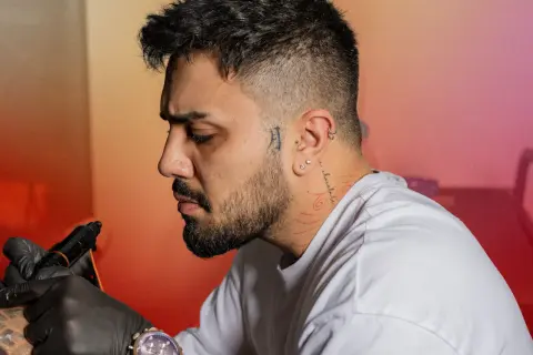 Un homme se fait tatouer le cou, un tatouage homme cou discret avec des motifs rouges. Il porte des gants noirs et une montre.
