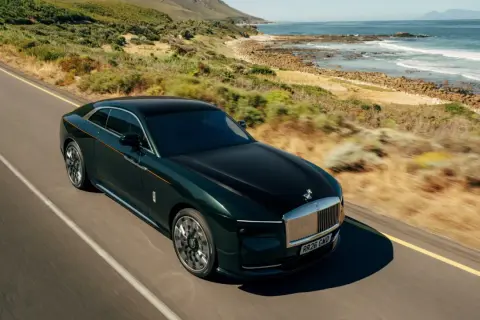 Elegancki, zielony Rolls Royce elektryczny sunie po malowniczej drodze nad oceanem.