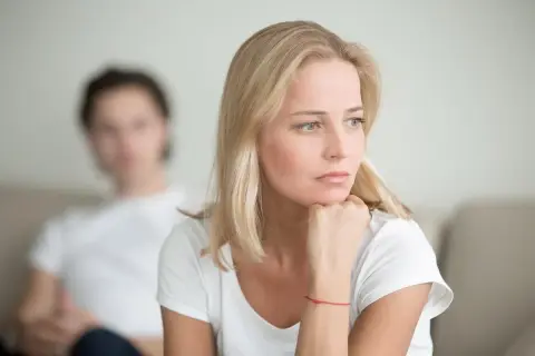 Zamyślona kobieta z czerwoną nitką na nadgarstku, zastanawia się, kto powinien odezwać się po rozstaniu. W tle niewyraźna postać mężczyzny.
