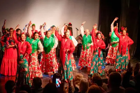 Un grupo de mujeres baila sevillanas de amor con trajes coloridos. Sus faldas amplias y mantones vibrantes llenan el escenario de alegría.