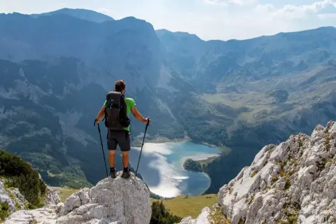 Trekking w Czarnogórze: turysta z plecakiem i kijkami stoi na skale, podziwiając górskie jezioro i pasma górskie.