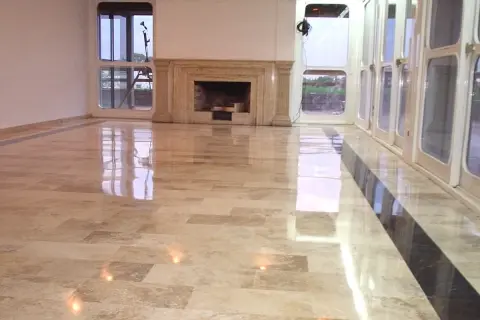 Pavimento in travertino lucidato a specchio, con un camino in marmo e grandi vetrate che riflettono la luce.