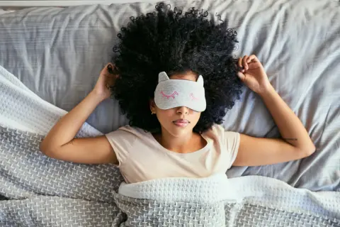 A woman with a large afro wears a cat-ear sleep mask, resting in bed. She's trying to figure out how long does it take to catch up on sleep.