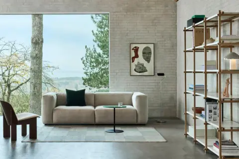 Gemütliches Wohnzimmer mit beigem Sofa, einem Holzsessel und einem Regal voller Bücher. Der Stil ist modern, mit einem Hauch von millennial grey in den Ziegelwänden.