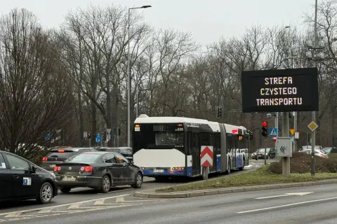 Autobus i samochody stoją w korku pod znakiem informującym o krakowskiej strefie czystego transportu.