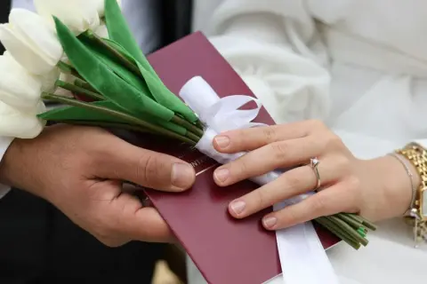 Dłoń mężczyzny i kobiety trzymają bukiet białych tulipanów i czerwony dokument.