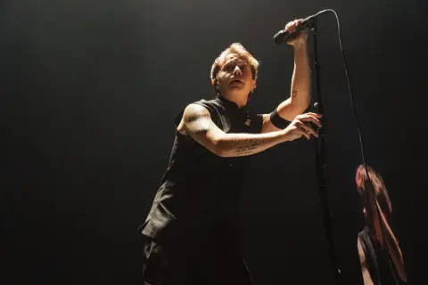 Lead singer of Nothing But Thieves, Conor Mason, belts into a microphone on a dark stage, his arm tattoos visible.