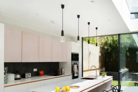 Cinq suspensions noires au design épuré, quel luminaire pour cuisine moderne ! Elles éclairent une cuisine lumineuse avec ses meubles roses et sa grande baie vitrée.