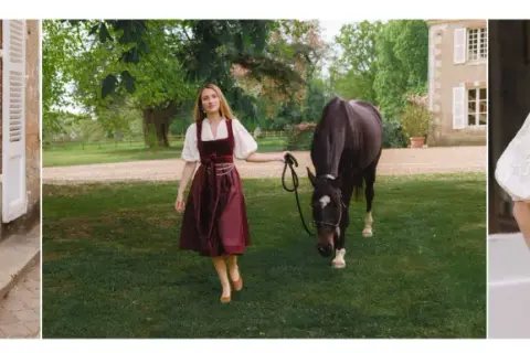 Eine Frau in einem weinroten Dirndl mit einer glänzenden Dirndlschürze aus Seide, daneben ein Pferd auf einer grünen Wiese.