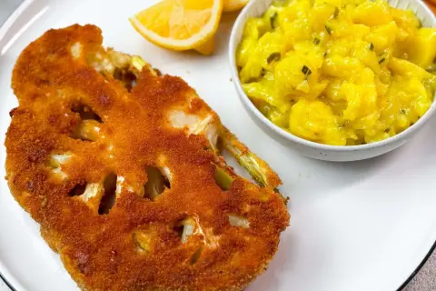 Knuspriges Blumenkohl-Schnitzel mit Kartoffelsalat und Zitronenspalten auf einem weißen Teller.