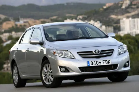 Srebrna Toyota Corolla 2010 z widocznym kierowcą, zaparkowana na tle krajobrazu z budynkami.