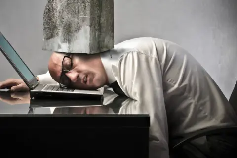 Man med sten på huvudet, utmattad vid sin laptop. Symboliserar digital stress och överväldigande arbetsbelastning.