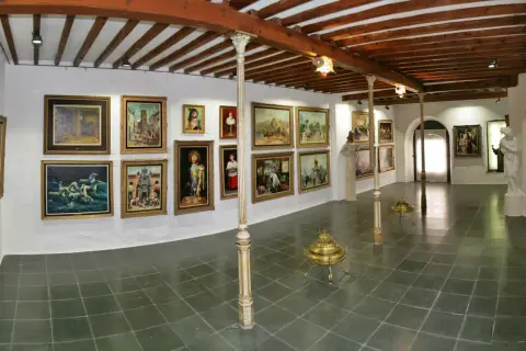 Galería de arte con cuadros y esculturas, techos de madera y suelo de baldosas.