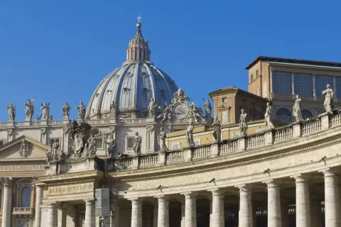 Cupola maestosa e colonnato di San Pietro, una delle quattro basiliche di Roma, con statue che vegliano sul cielo azzurro.