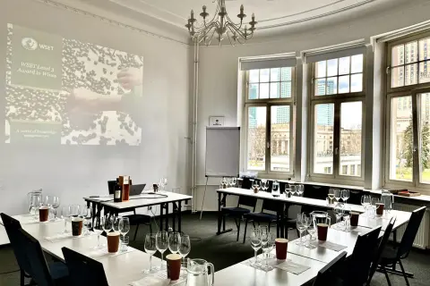 Sala do degustacji win, przygotowana do spotkania klubu wino. Na stołach kieliszki, dzbanki z wodą i butelka wina. Na ekranie logo WSET.
