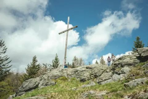 Wanderer auf einem felsigen Gipfel im Bayerischen Wald unter einem Holzkreuz. Ein typischer 8-Tausender-Ausblick.