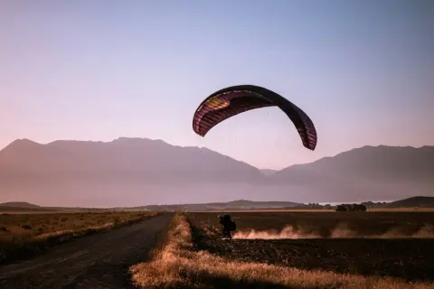 Parapentista aterrizando en un campo polvoriento al atardecer, rodeado de montañas. Una escena de turismo activo.