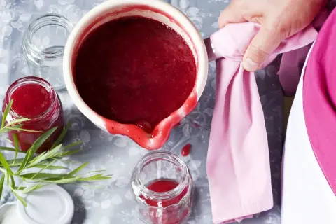 Frisch gekochte, einfache Johannisbeermarmelade wird in ein Glas gefüllt. Ein Hauch von Grün und ein rosa Tuch vervollständigen das Bild.