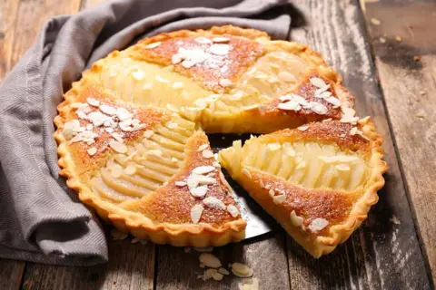 Une délicieuse tarte aux poires, garnie d'amandes effilées et saupoudrée de sucre glace, repose sur une table en bois.