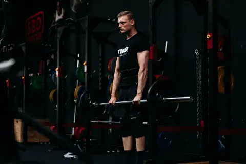 Ein Mann mit Bart und schwarzem T-Shirt hebt eine Langhantel im Fitnessstudio. Er trainiert seine Handkraft, um stärker zu werden.