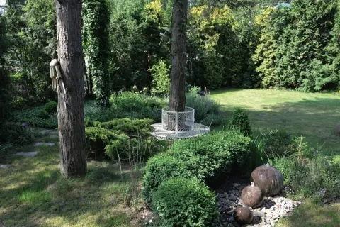 Urokliwy ogród w lesie z kamiennymi kulami tworzącymi wodną kaskadę, ozdobną ławką wokół drzewa i bujną zielenią.