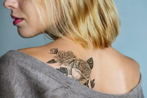 Ein frisches Tattoo mit Rosen auf der Schulter einer Frau, das mit einem grauen Pullover abgedeckt wird.