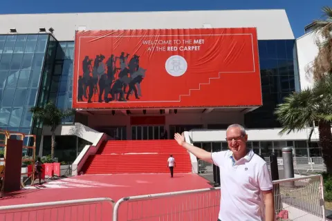 Mężczyzna pozuje przed budynkiem z wielkim czerwonym banerem "Welcome to the Met at the Red Carpet". To przygotowania do festiwalu w Cannes.