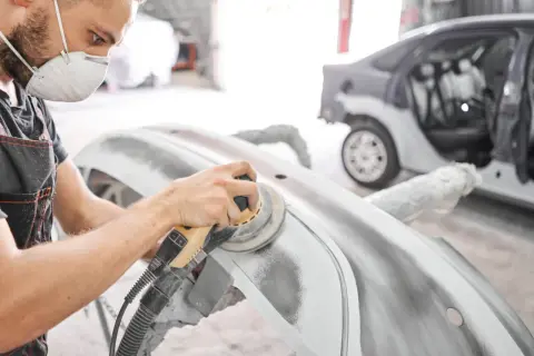 Pracownik w masce ochronnej wykonuje szlifowanie szpachli samochodowej na zderzaku. W tle widać auto w trakcie naprawy.