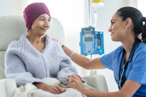 Eine Krankenschwester tröstet eine Patientin während des Chemotherapieablaufs. Die Patientin trägt eine lila Kopfbedeckung und eine Strickjacke.