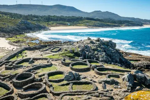 Ruinas circulares de piedra del castro de Baroña, con el mar azul y una playa de arena al fondo.