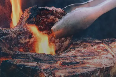 En saftig entrecote grillas över öppen eld, perfekt innertemp. Tången vänder köttet för en jämn stekyta.