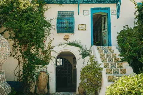 Fachadas antiguas con escaleras de azulejos coloridos, puerta azul y vegetación exuberante. Un portal arqueado y vasijas de barro completan la escena.