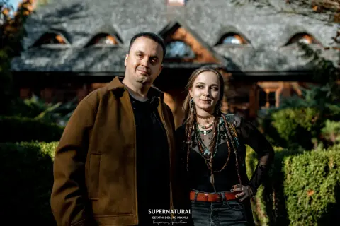 Para pozuje przed domem z kamiennym dachem. Mężczyzna w brązowej kurtce i kobieta w czarnej bluzce z wieloma naszyjnikami. Wyglądają jakby wyszli z planu filmu o straszydłach.