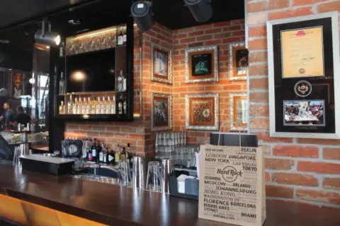 Wnętrze Hard Rock Cafe w Gdańsku: bar z butelkami alkoholu, ozdobne ramki na ścianie z cegły i torba z nazwami miast.