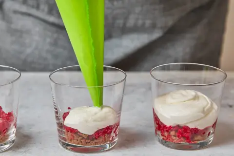 Cremige Masse wird mit Spritzbeutel in Gläser mit Himbeeren und Spekulatius-Bröseln gefüllt.